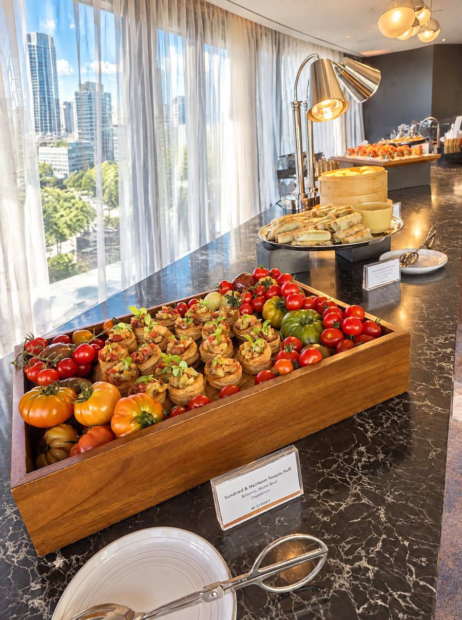 W Sydney, buffet counter with a timber tray of heirloom tomatoes and tomato puffs, heat lamps and a bamboo steamer behind, floor to ceiling windows onto city towers and trees